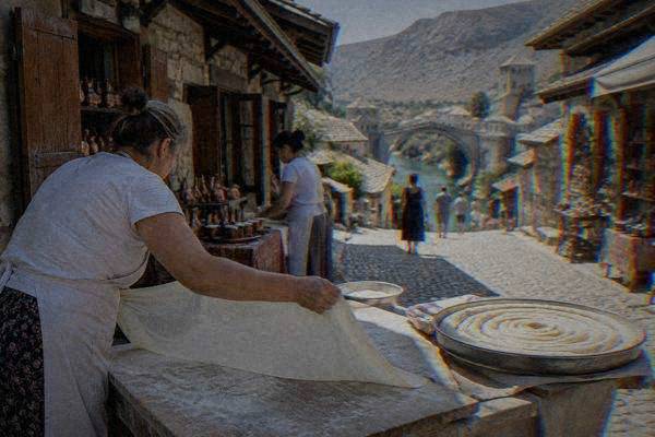 Hands stretching a paper-thin sheet of dough across a floured table, a tray of coiled pastry beside it, a few people in aprons, Kujundziluk